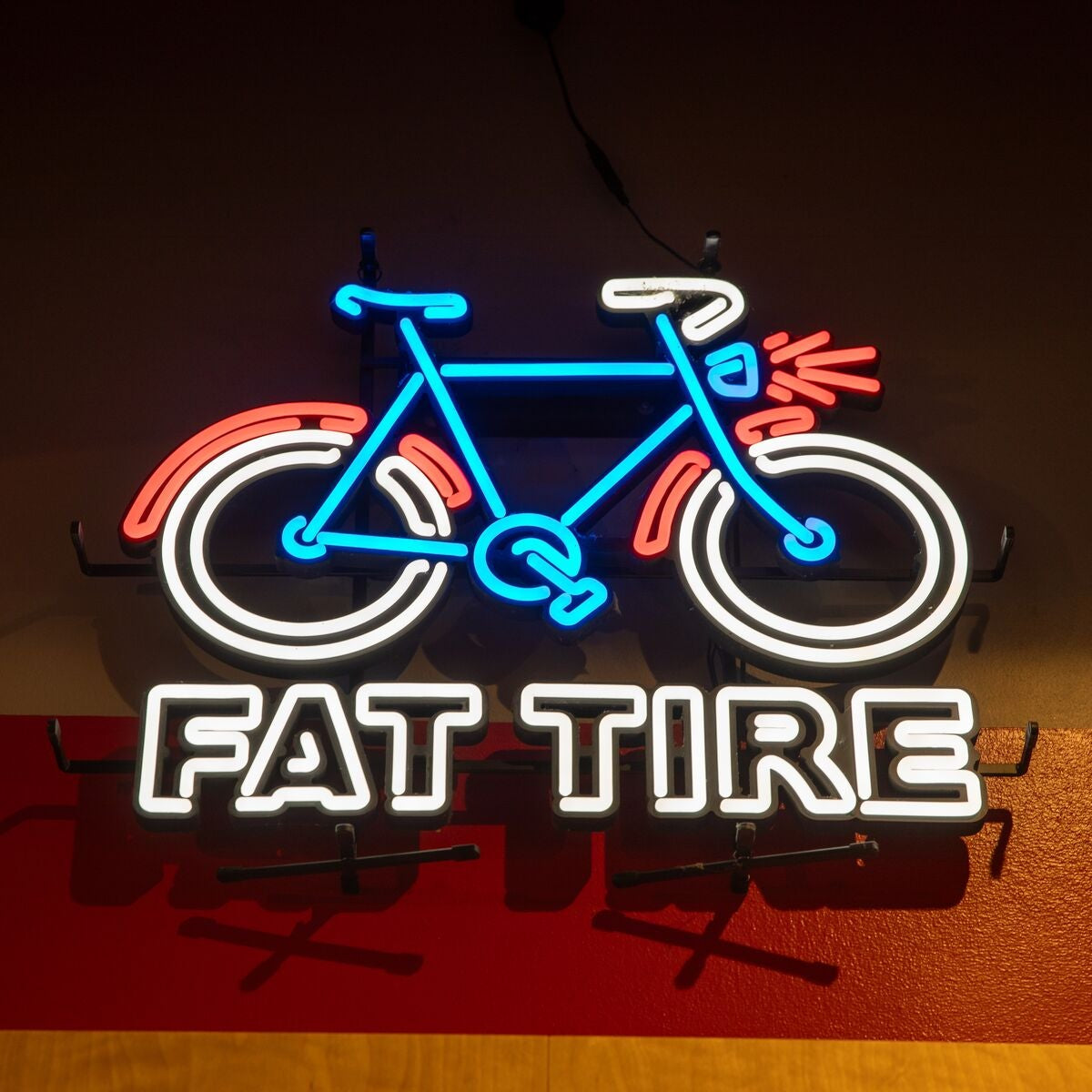 A neon sign featuring a bicycle with the words 'FAT TIRE' in glowing white letters below it, mounted against a dark wall with a red horizontal panel.